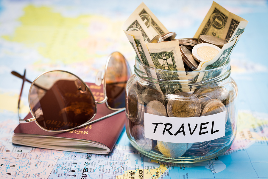  A photo of a jar filled with money, a passport with sunglasses on top of it, and a map in the background with the word 'TRAVEL' on a tag around the jar.